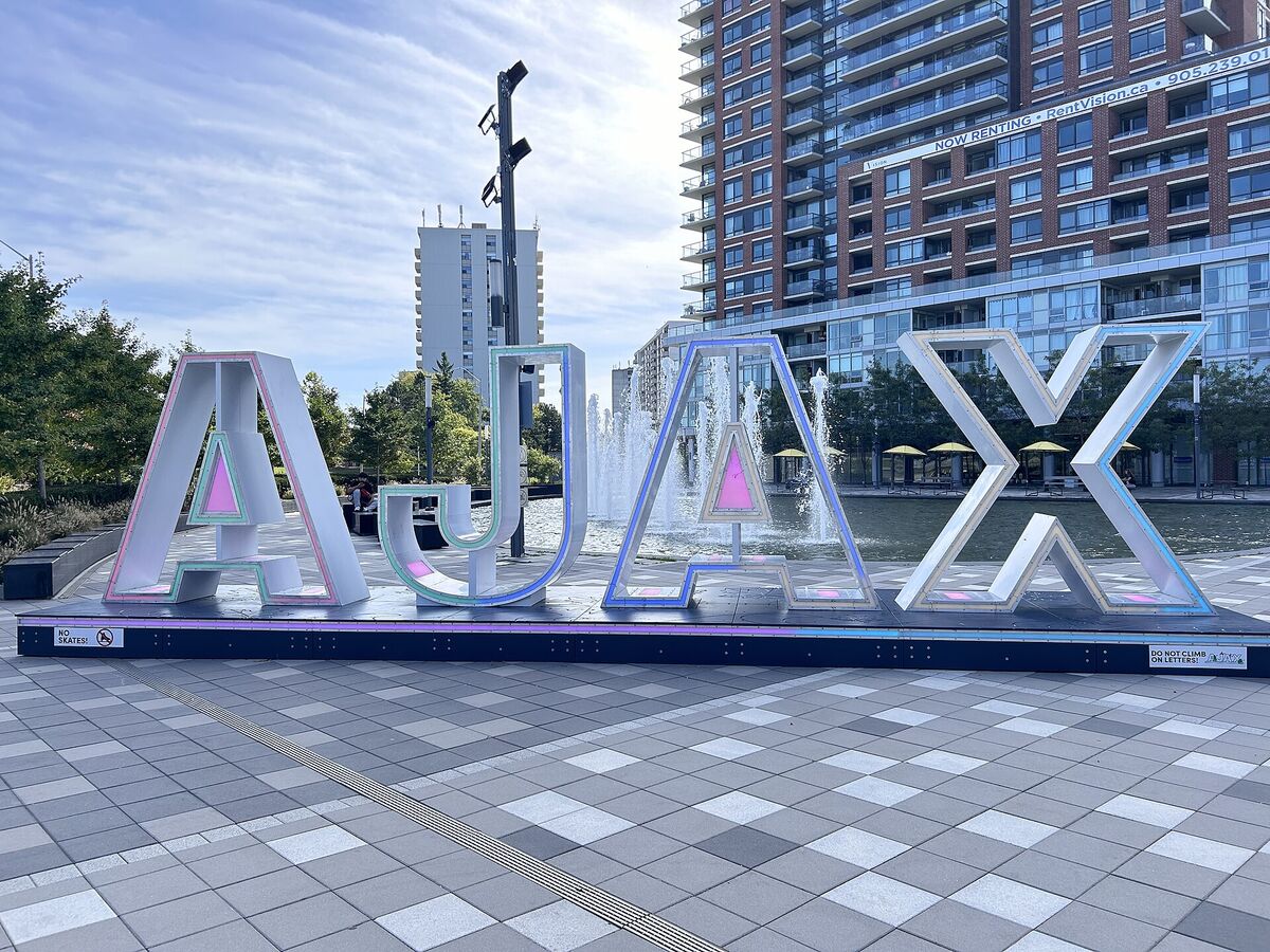 Large illuminated letters spelling AJAX in a public square with fountains and apartment towers